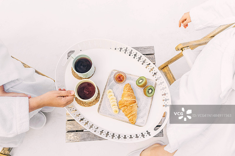 两位美丽的年轻女子穿着白色毛巾浴袍在酒店享用早餐，欣赏美丽的山景图片素材