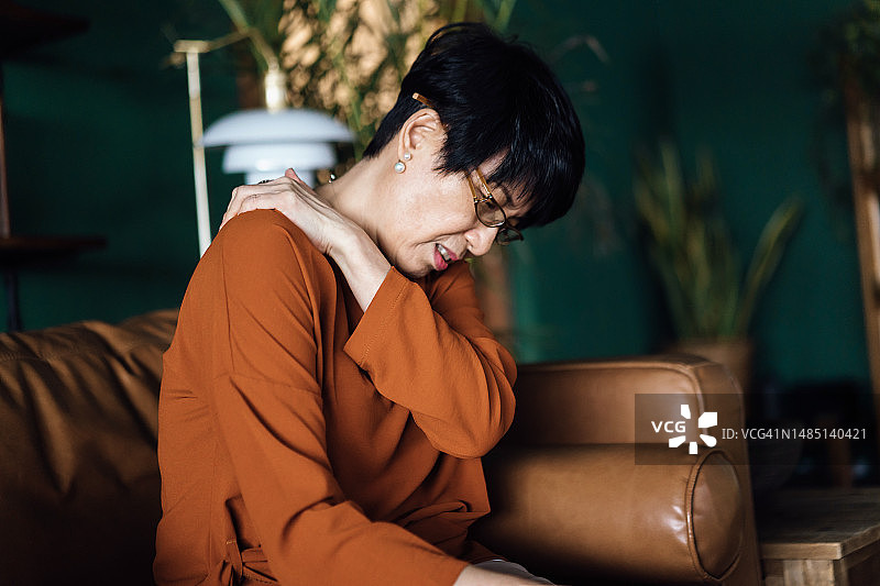 一位心烦意乱的亚洲老年妇女感到不适，颈肩疼痛，坐在家里的沙发上按摩酸痛的肌肉。老年人和健康问题概念图片素材