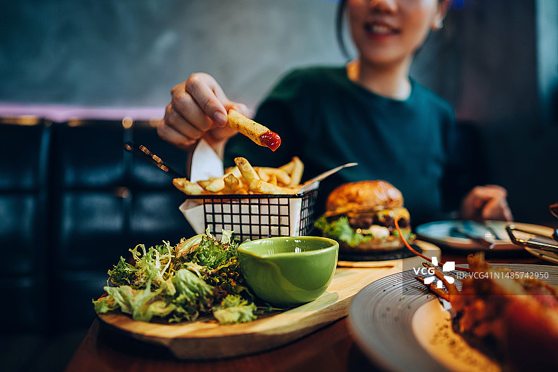 年轻的亚洲女性吃着新鲜制作的美味汉堡，在餐桌上将炸薯条蘸番茄酱的裁剪照片。在餐厅享受盛宴。人物、食物和生活方式图片素材