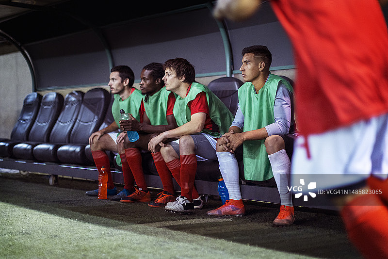 专业足球运动员在替补席观看队友比赛的侧面视角图片素材