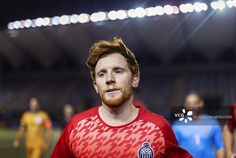 在大型比赛前站在体育场灯光照亮的球场上的红发专业男足球运动员的肖像图片素材