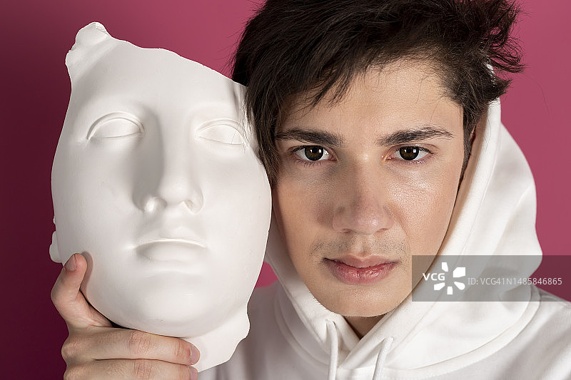 身穿白色连帽衫，用面具遮住脸的可疑年轻人画像。秘密崇拜者，隐藏身份概念。粉色背景隔离图片素材