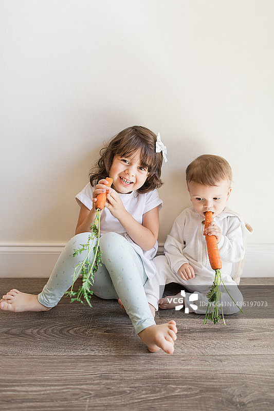 2023年春季，一个4岁的古巴裔美国卷发女孩在家中简约空间里赤脚吃着胡萝卜，旁边是她穿着白色兔子服装的15个月大的弟弟图片素材