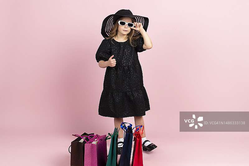 购物的孩子。迷人的小女孩身穿黑色时尚夏装，戴着遮阳帽和酷炫的白色太阳镜，化着淡妆，摆着购物袋的造型。粉红色背景上孤立。图片素材