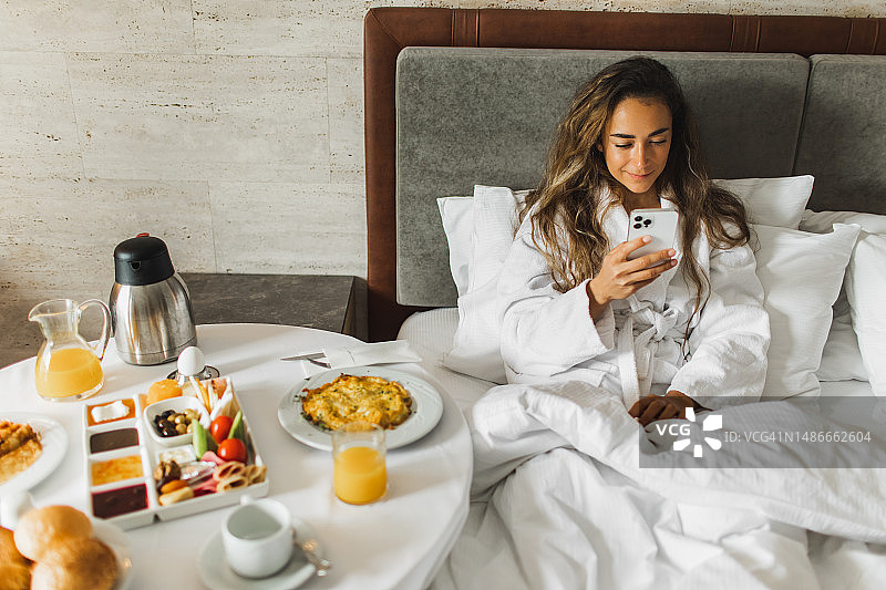 女人躺在床上玩手机，桌上放着酒店早餐图片素材