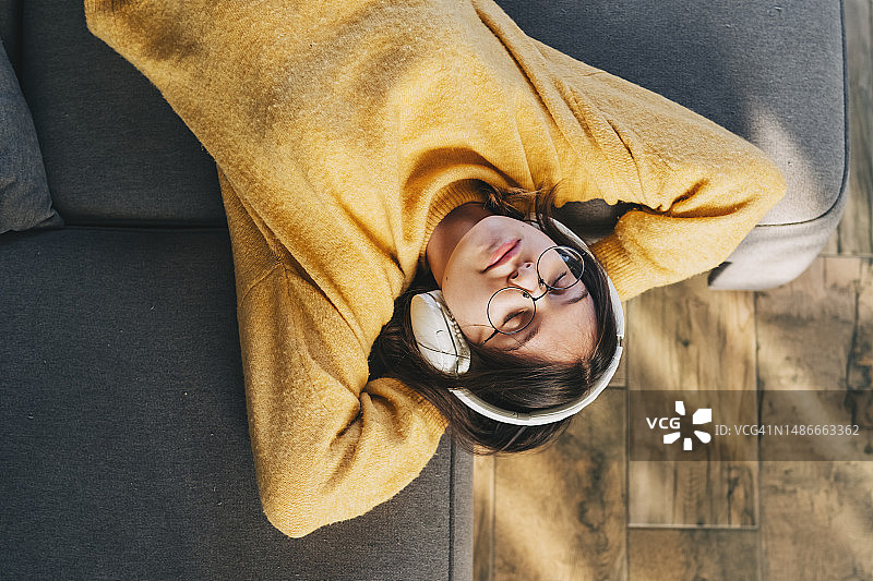 快乐的青少年女孩和猫在沙发上，年轻女人戴着耳机听音乐图片素材