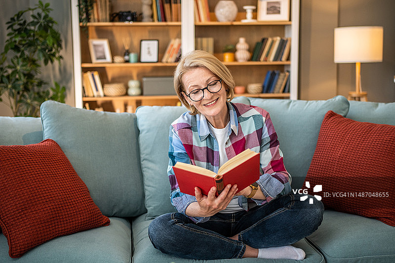 快乐的中年妇女坐在沙发上读书图片素材