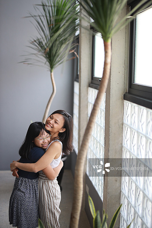 站在客厅窗边，亚洲妈妈亲切地拥抱女儿，展现爱、关怀和家庭亲情图片素材