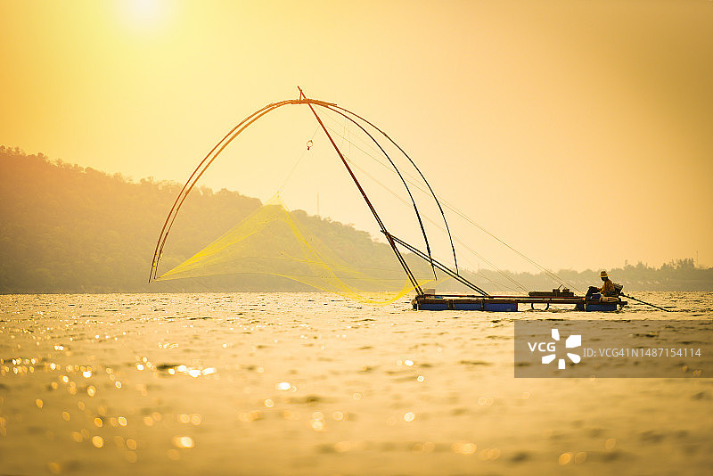 亚洲渔民在木船上用渔网捕鱼，在河中撒网，日落或日出——轮廓渔船，背景是山岛，在海洋上，罗马尼亚图片素材