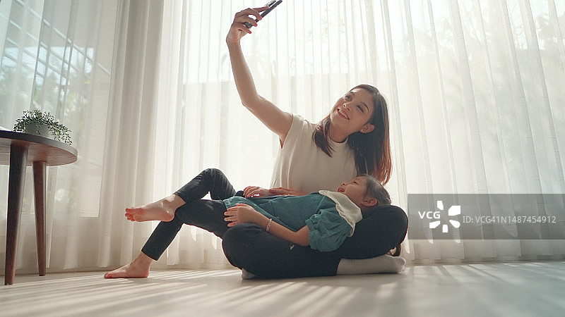 年轻的亚洲妈妈和可爱的女儿舒适地坐在家里，拥抱在一起，一起拍照。育儿和对儿童成长和良好发展的关注，快乐的母性时刻。家庭概念。图片素材