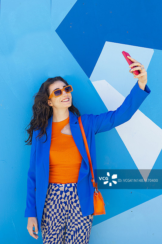 快乐的戴着太阳镜在蓝色墙壁前自拍的女人图片素材