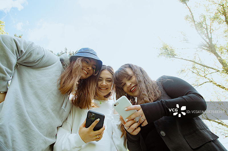 白天三位拿着手机微笑的棕色长发女性朋友仰视角度图片素材