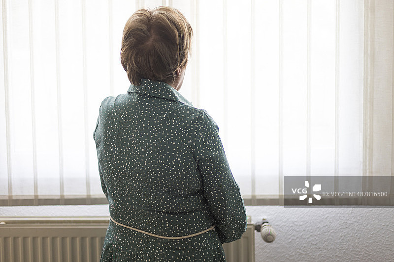 患有阿尔茨海默症和抑郁症的年长女士，背对着窗户，若有所思地向外看。关于老年、疾病、孤独、健忘和记忆的概念。图片素材