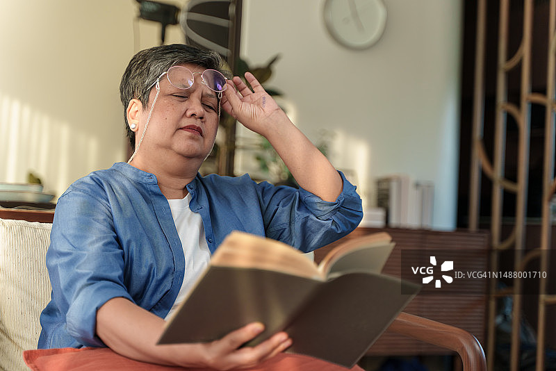 患有远视老花眼、摘下眼镜看书的年长亚洲女性肖像：视力不佳，有远视问题，老年健康状况问题图片素材