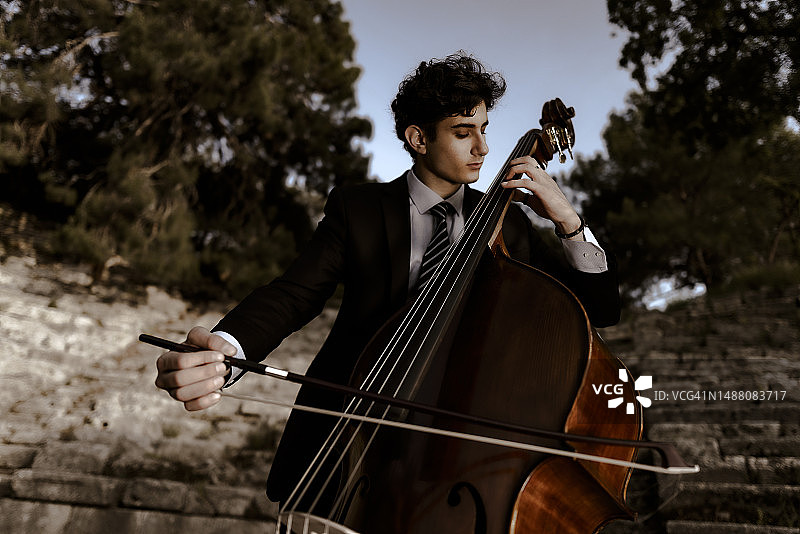 身穿黑色西装的年轻音乐家在历史悠久的竞技场和大自然中演奏低音提琴图片素材