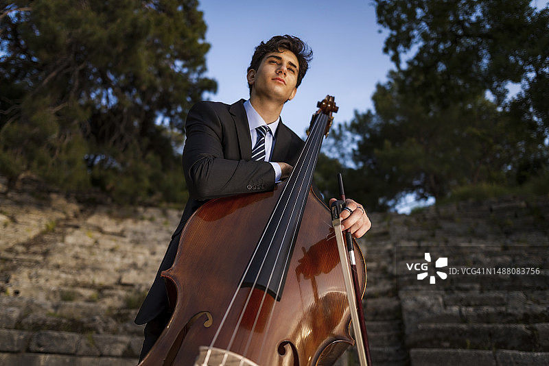 身穿黑色西装的年轻音乐家在历史悠久的竞技场和大自然中演奏低音提琴图片素材
