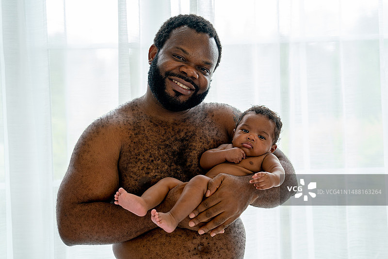 没穿衬衫的非裔美国父亲抱着新生婴儿站在玻璃窗前，父亲看着镜头微笑图片素材