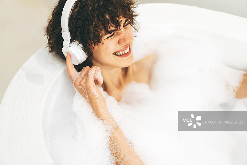 漂亮的女人在泡泡浴中进行自我约会，戴着耳机听音乐图片素材