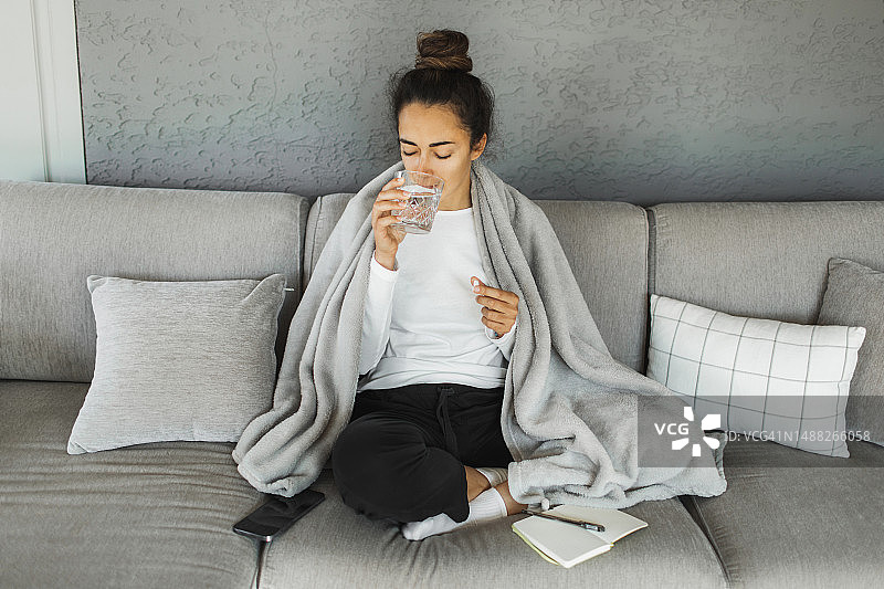 发烧坐在沙发上，盖着格子呢毯子并服用退烧药的女人图片素材