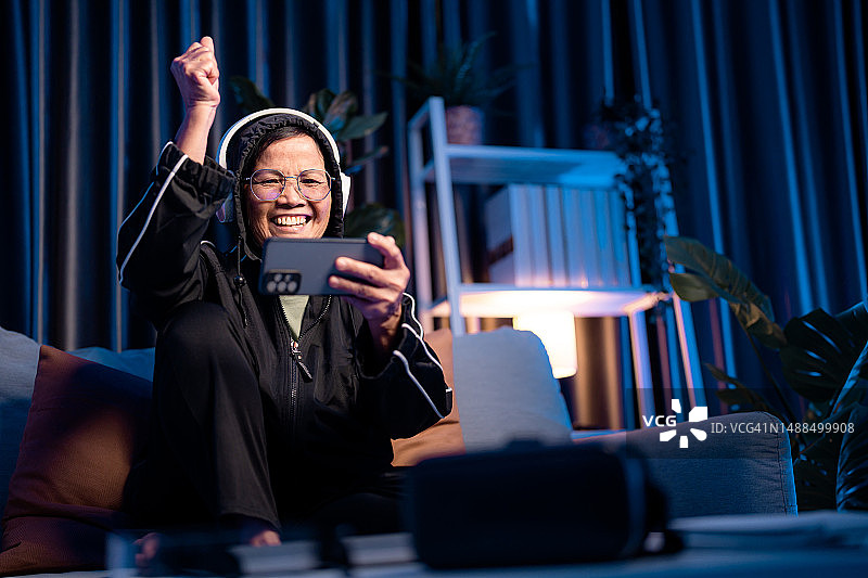 年长的亚洲女人戴着耳机在沙发上玩手机游戏图片素材