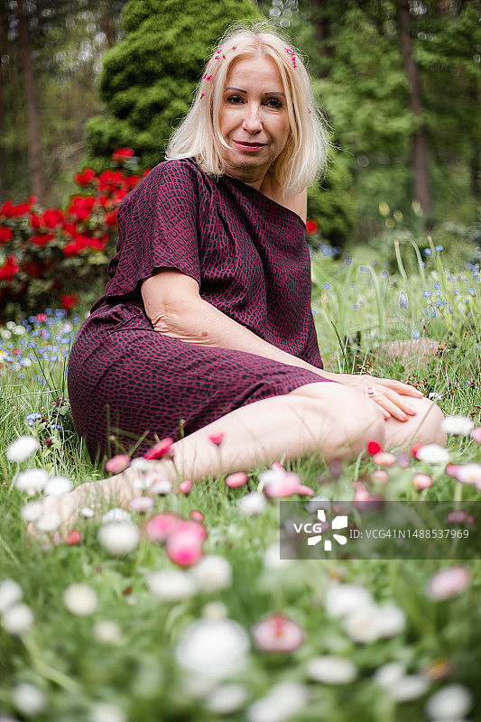 身穿深紫色连衣裙的漂亮女人，坐在绿草地上，背景是红色鲜花和森林图片素材