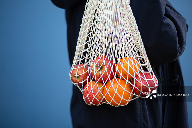 女人拿着装满水果的可重复使用网袋，背景为蓝色图片素材