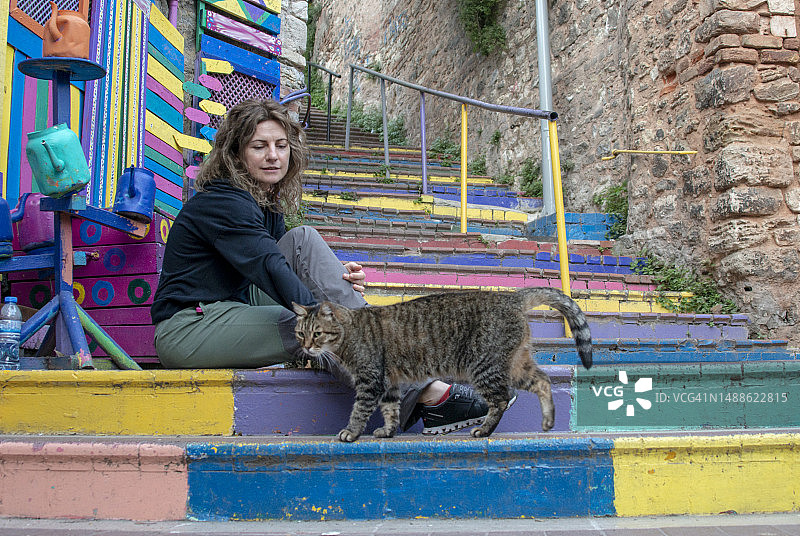 年轻女人坐在土耳其伊斯坦布尔巴拉特区的楼梯上，和可爱的猫玩耍图片素材