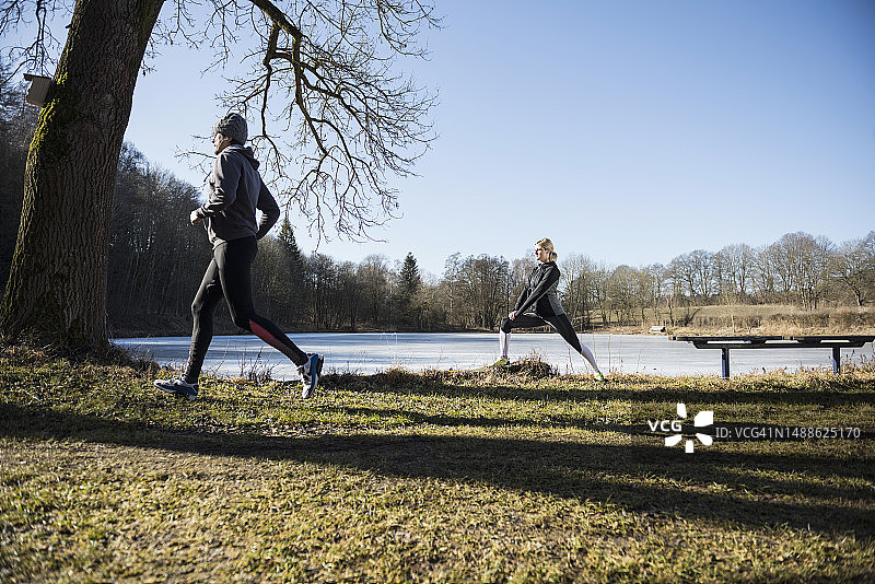 在冰冻的湖前健身步道上慢跑的男人和伸展身体的女人图片素材