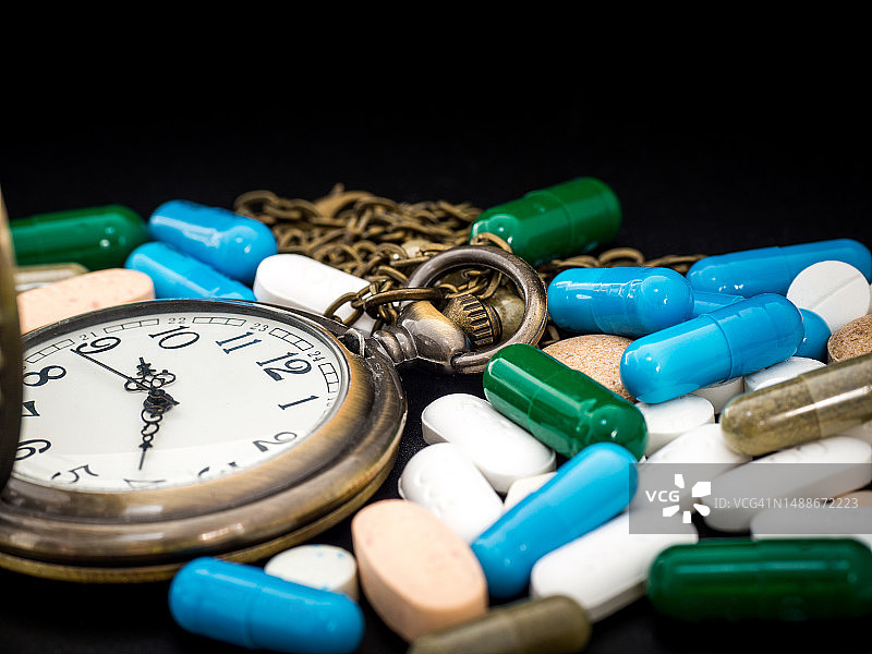 黑色背景上的古董时钟与药物胶囊特写：反对药物滥用，绿色蓝色白色圆形胶囊药丸图片素材