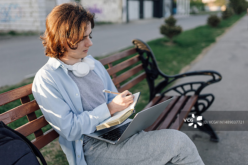 年轻作家在公园长椅上用笔记本电脑写作的侧视图图片素材