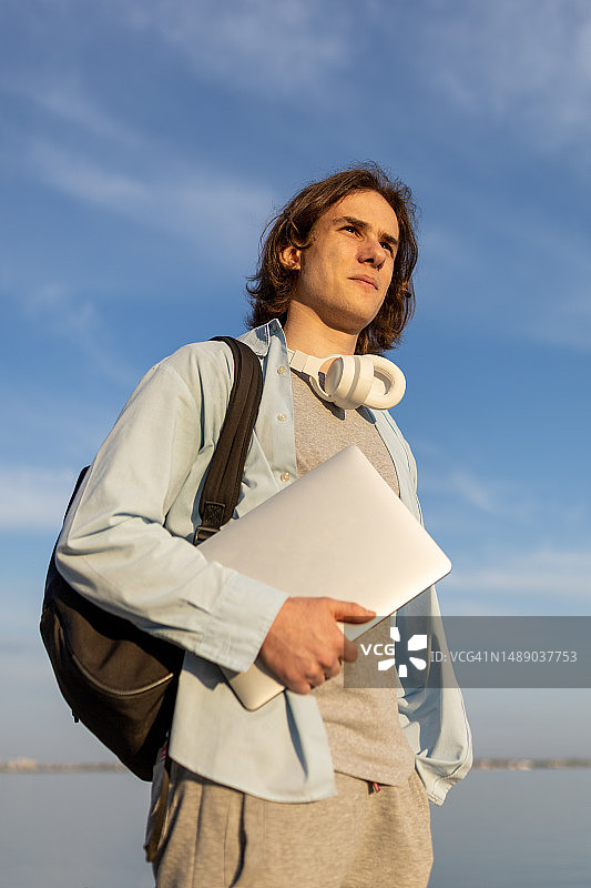 傍晚阳光下，手持笔记本电脑和黑色背包的年轻男子图片素材