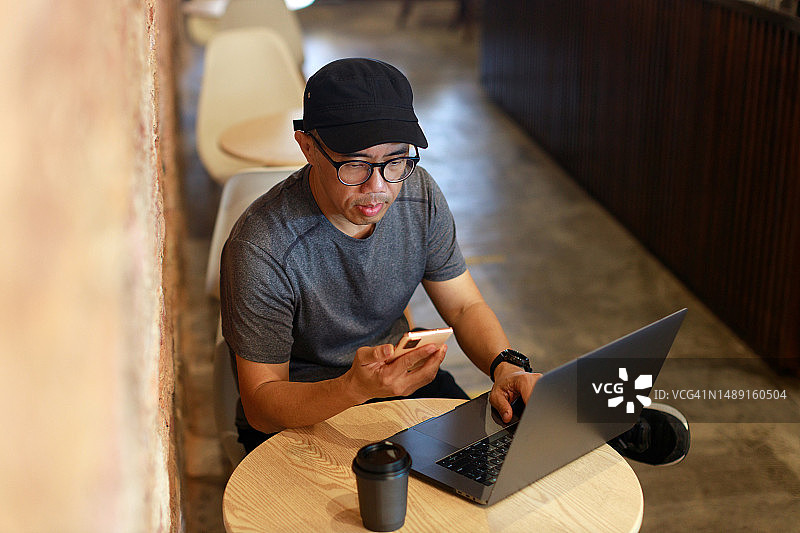 戴着帽子的亚洲男子坐在咖啡馆里，一边喝咖啡一边用电脑和智能手机处理多项任务图片素材