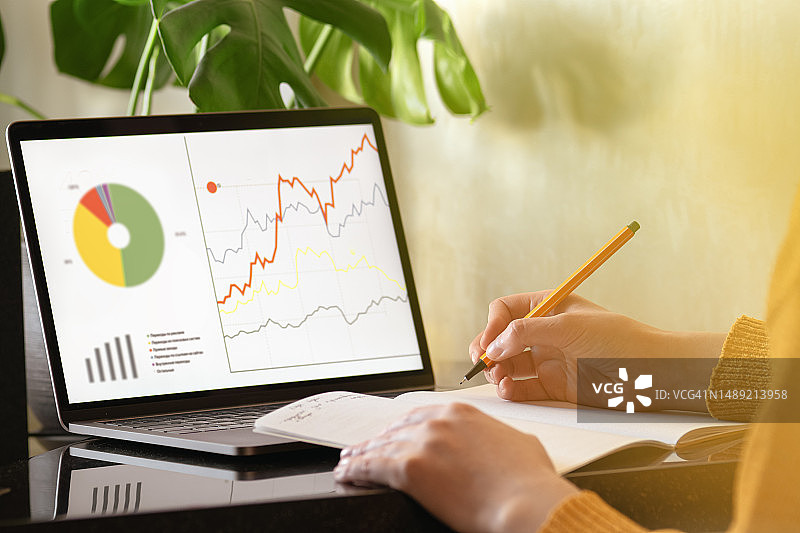 穿着黄色开衫的女人记录笔记本电脑屏幕上的数字，交易分析、业务增长、营销、数据、统计、安全纸、银行概念图片素材