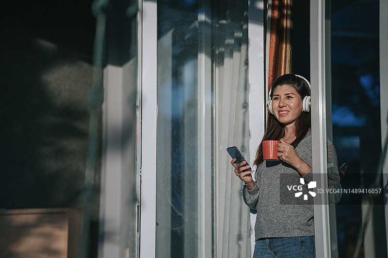 一名亚裔华裔女子在日落时分倚在滑动门上用智能手机听音乐图片素材