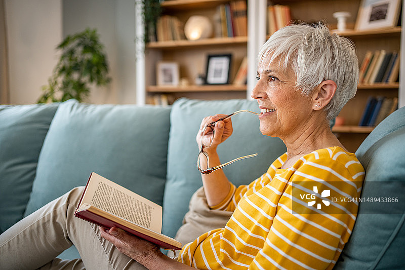一位退休妇女舒适地坐在公寓的沙发上，专心阅读她最喜欢的书图片素材