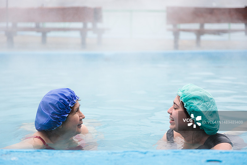 拉丁家庭的宁静：老妇和年轻女儿身穿泳衣和浴帽，在雾气弥漫的温泉池中聊天图片素材