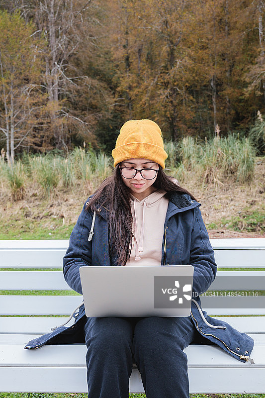 在自然环境中远程工作：一位穿着暖和的衣服并戴着眼镜的年轻女子，在森林中一个宁静的长椅上用笔记本电脑远程办公的正面图。图片素材