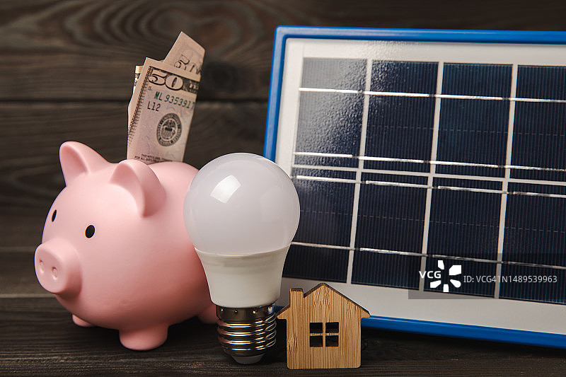 棕色木头上的太阳能电池板、LED灯、房屋模型、钱和储蓄罐扁平风格构图，节约金钱和清洁能源的概念图片素材