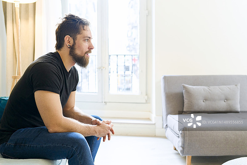 穿休闲装、留着胡须的男人沉思着坐在客厅沙发上（侧面）图片素材