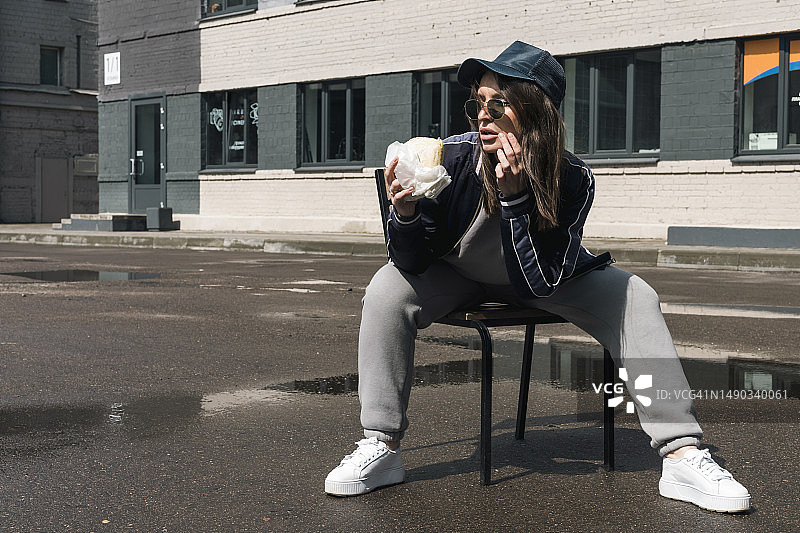 年轻的黑发女孩穿着休闲运动服和太阳镜在街上吃快餐汉堡图片素材