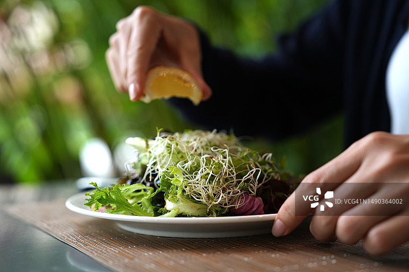 女子将柠檬汁挤在沙拉上的局部照片，沙拉配有苜蓿芽、樱桃番茄、核桃、绿叶蔬菜、橄榄和沙拉酱。图片素材