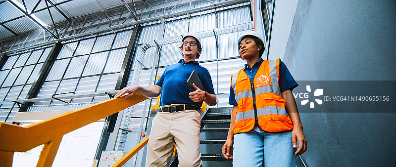 中年白人男性企业家经理与黑人女工程师技术员在工厂合作，交谈并走下楼梯。物流行业业务、工业工作或工作中的人们图片素材