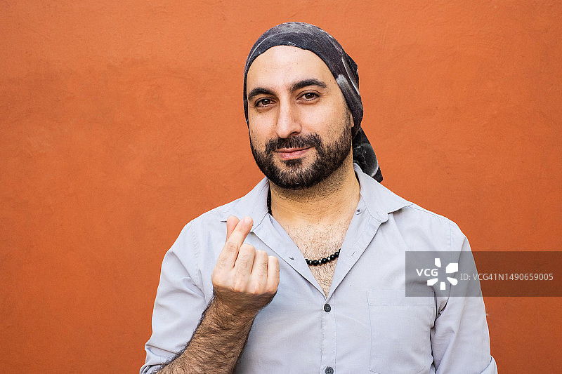 戴着头巾的中东裔拉丁美洲男人面带微笑，看着镜头，用食指和拇指做出韩国爱心手势图片素材