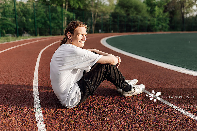侧视角度：年轻跑步者坐在红色跑道上，抱着膝盖微笑图片素材