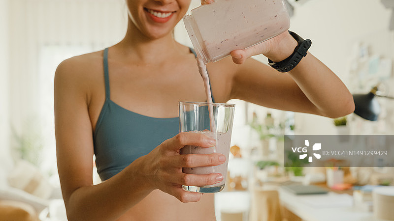 年轻亚洲女性在家使用搅拌机并将冰沙倒入玻璃杯中，健康食品图片素材