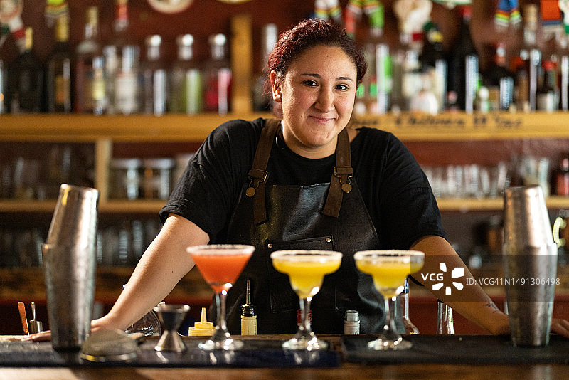 女调酒师在吧台后调制彩色鸡尾酒，对着镜头微笑图片素材