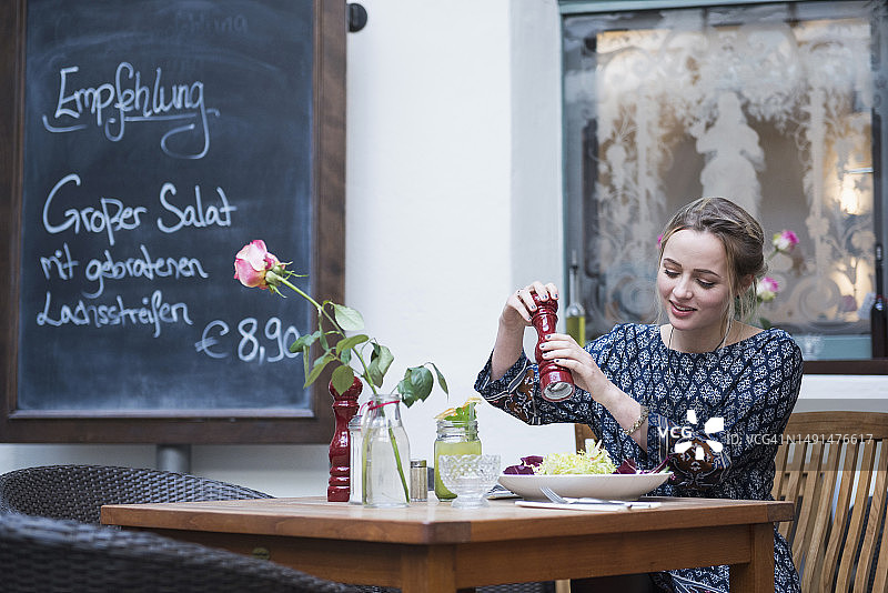 年轻女人在咖啡馆的黑板旁把胡椒粉撒在沙拉上图片素材