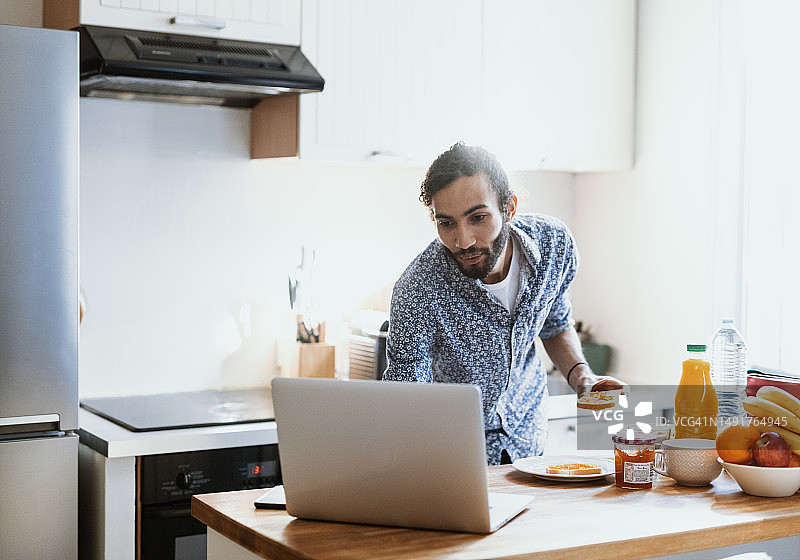 在家准备早餐并在厨房岛台上使用笔记本电脑的中年混血男人图片素材
