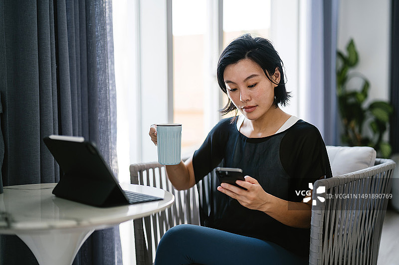 年轻的亚洲女人坐在窗边的桌子旁，查看她的智能手机并在家中使用平板电脑。管理银行和金融。网上银行概念图片素材