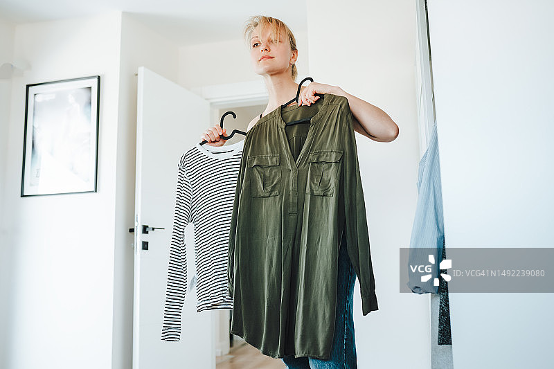 购物狂的烦恼：选择服装搭配，明亮客厅里手拿衬衫的女人图片素材